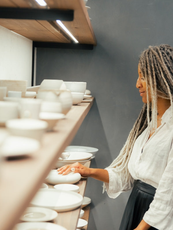 A woman with dreadlocks shops for ceramics, thoughtfully touching a plate on a wooden shelf, evoking a curated and globally-inspired home.
