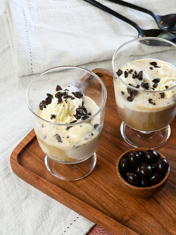 Two glasses of affogato on a wooden tray, topped with chocolate shavings and served with chocolate-covered espresso beans, creating a rich and inviting dessert scene.
