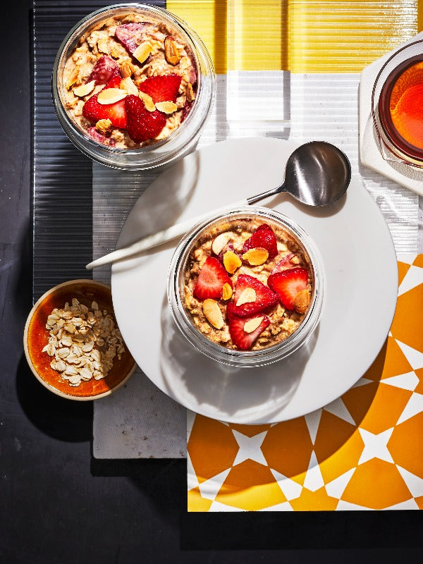 Overhead shot of two glass jars of strawberry overnight oats, arranged artfully with a spoon, dish of oats, and colorful backgrounds.
