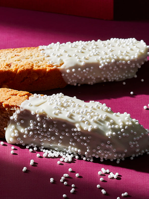 Two almond biscotti dipped in white chocolate and sprinkles sit invitingly on a magenta surface. A tempting treat!
