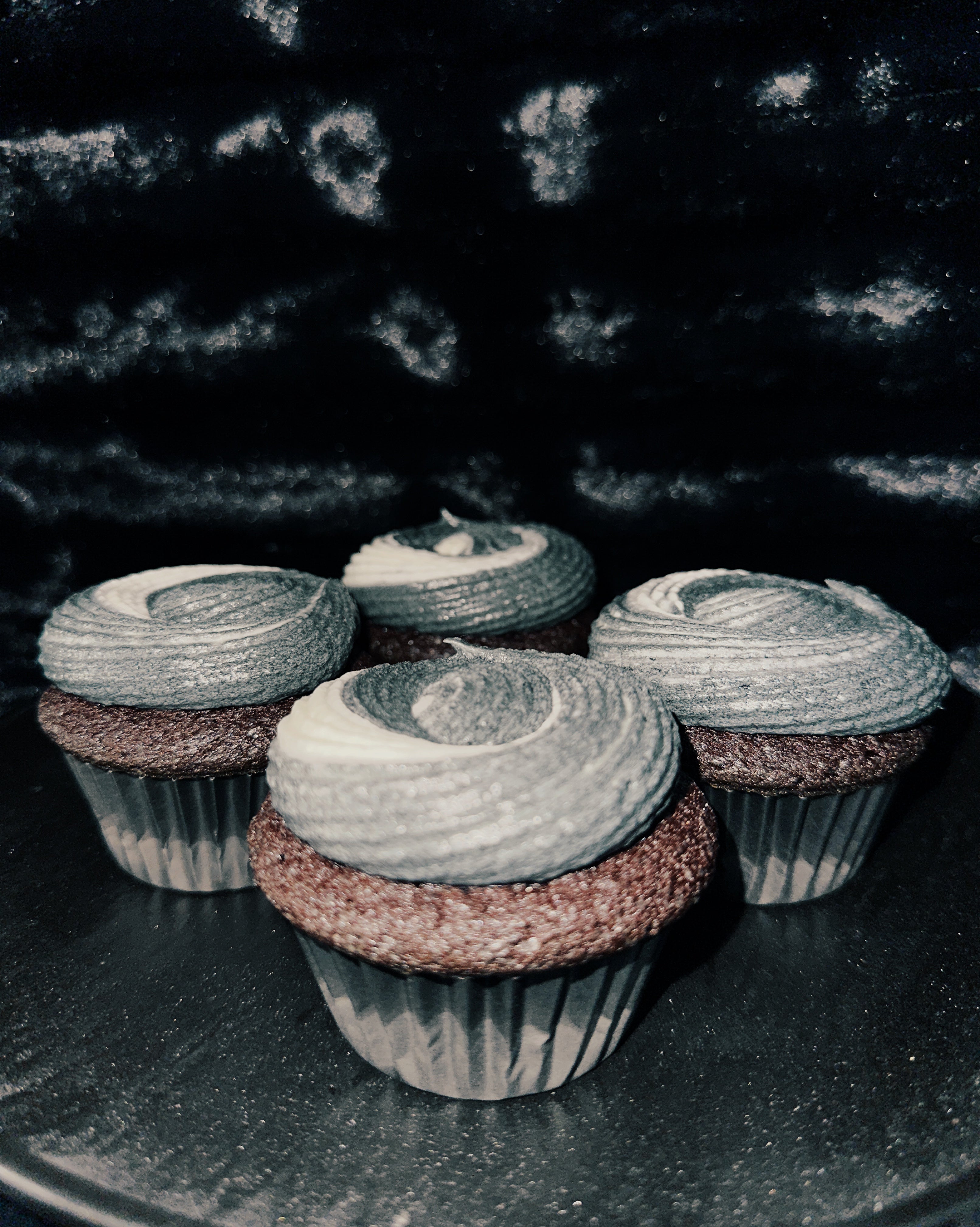 Moody shot of McBay's black velvet cupcakes featuring dark gray frosting on a glittery black surface, creating an eerie, elegant feel.
