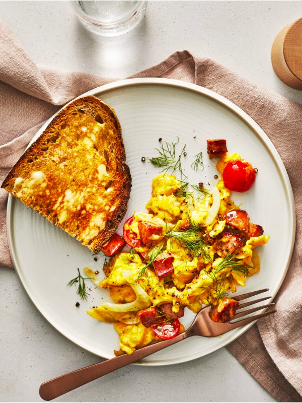 A light and airy breakfast scene of scrambled eggs with chicken sausage, fennel, and buttered toast on a plate. 
