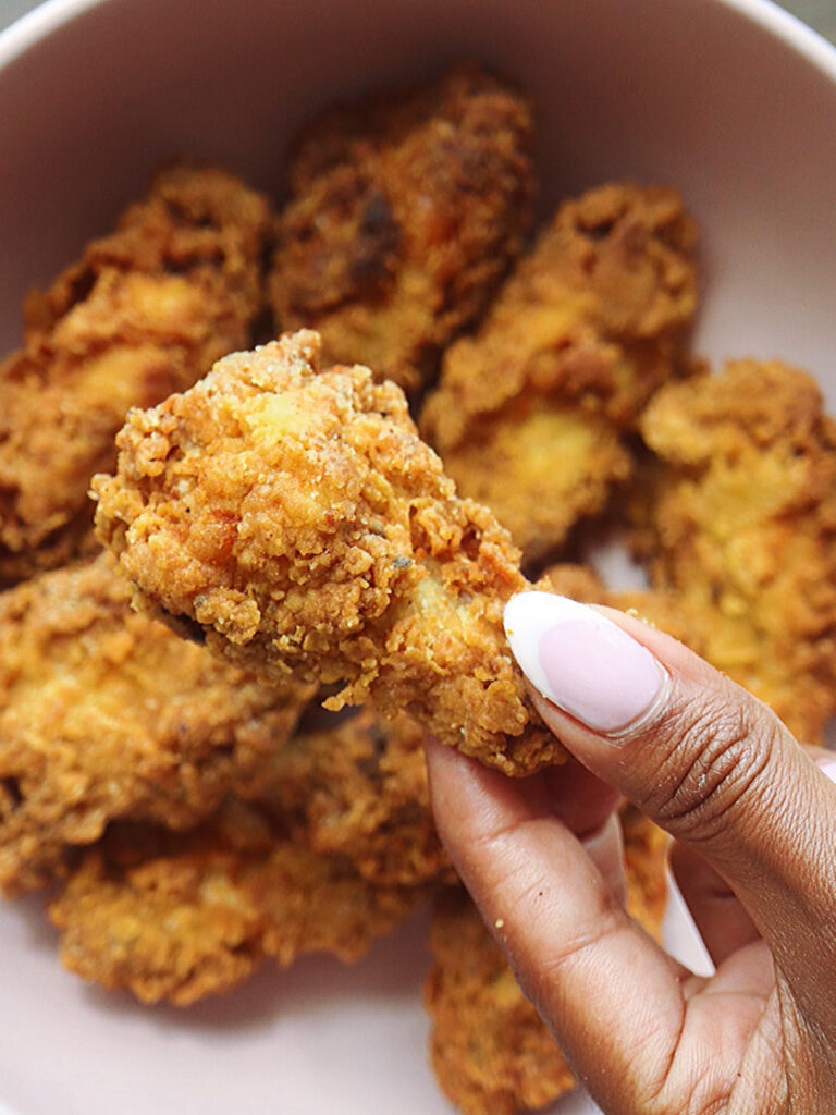 A crispy fried chicken leg, held by a hand with a pink and white manicure, sits above a bowl of more chicken.