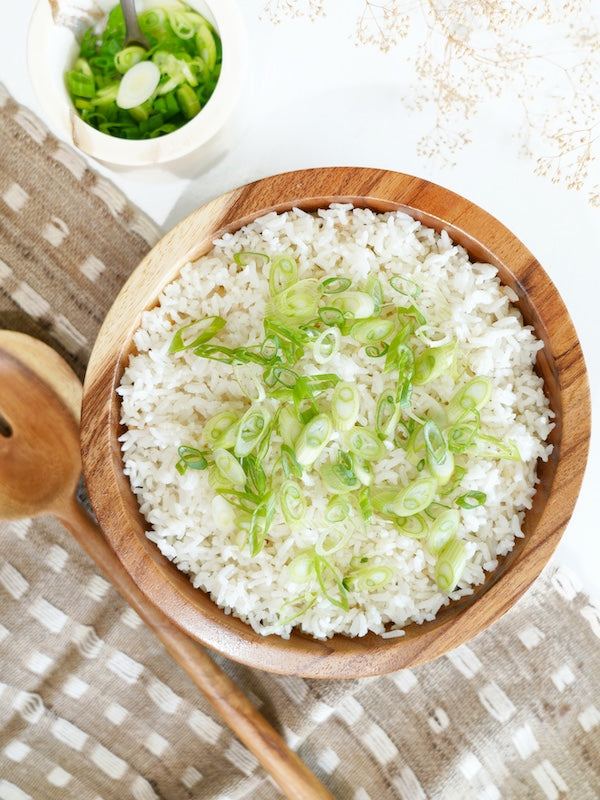 Overhead shot of fluffy coconut rice topped with fresh green onions, served in a wooden bowl on a patterned cloth. 
