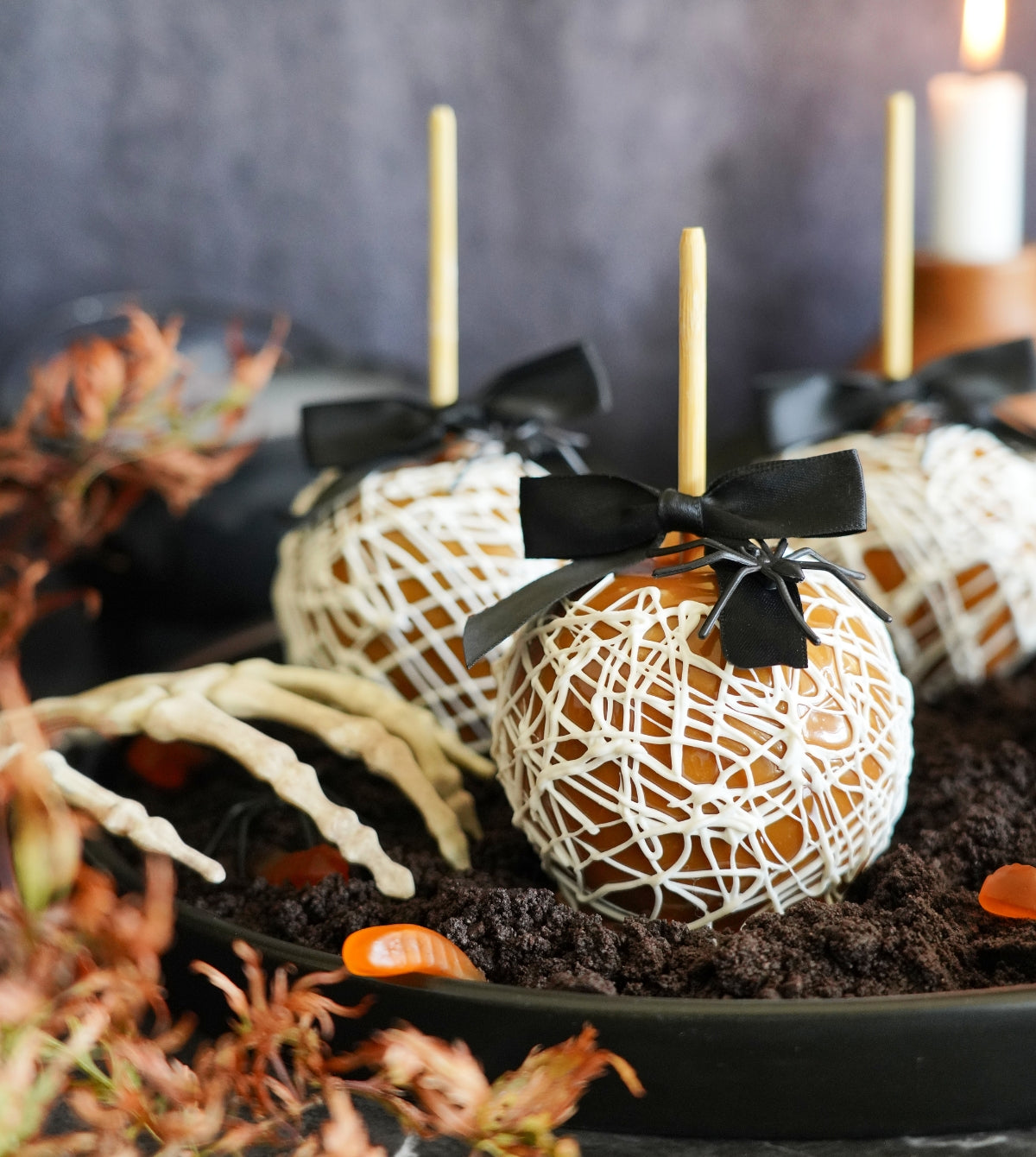 Spooky caramel apples, adorned with white chocolate webs and black bows, sit in "dirt" alongside a skeleton hand for a Halloween treat.

