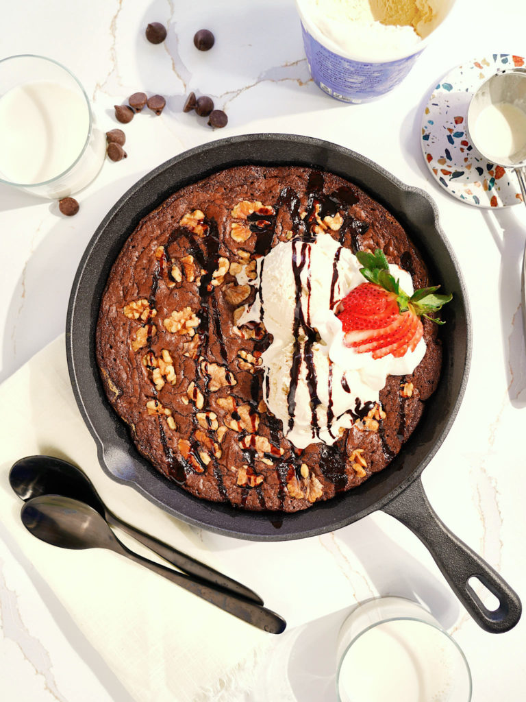 Warm dark chocolate skillet brownie, topped with vanilla ice cream, strawberries, walnuts, and chocolate syrup, next to a glass of milk.
