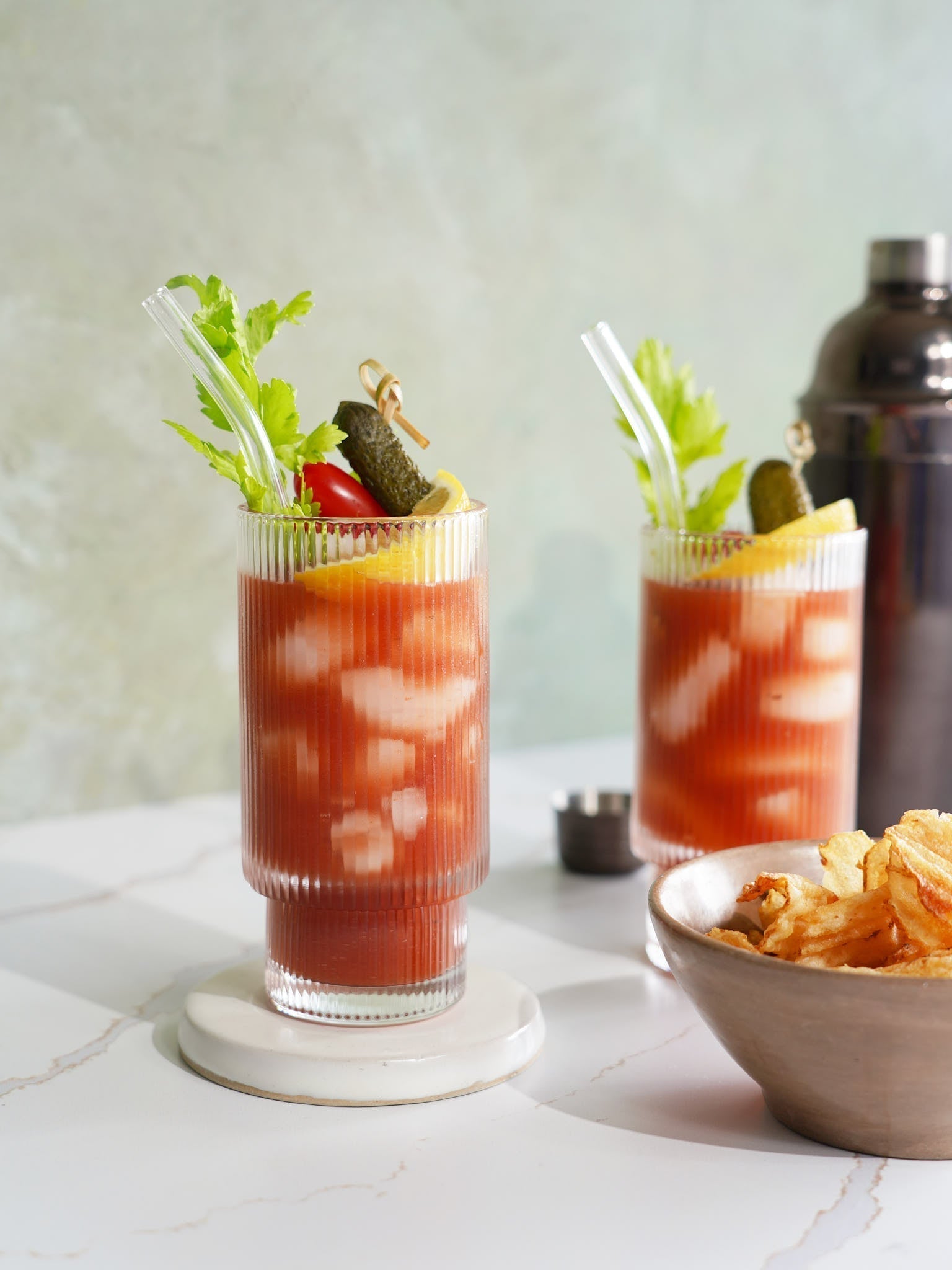 Two Bloody Maria cocktails garnished with celery, lemon, and a pickle, alongside a bowl of chips, set against a modern backdrop.
