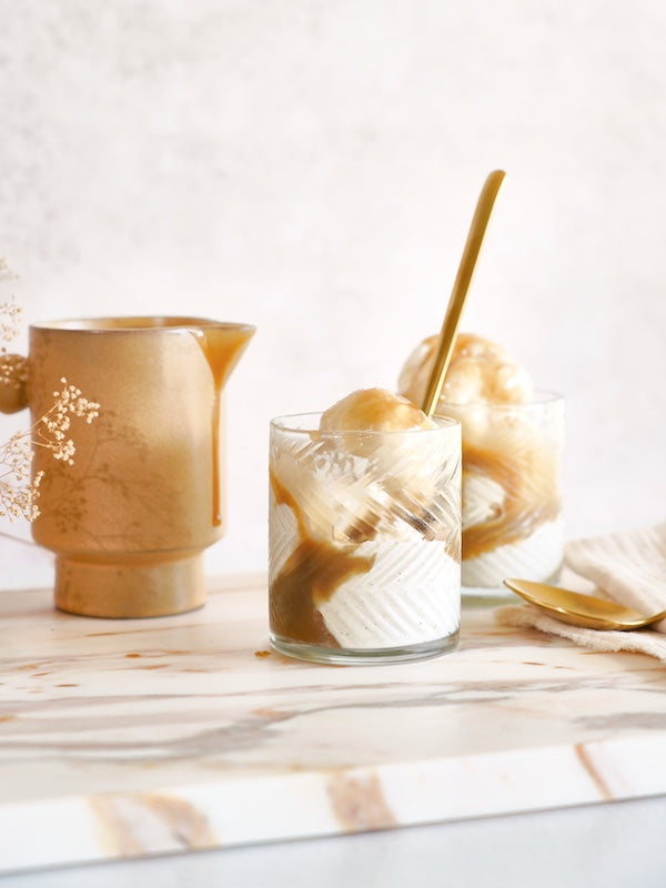 Two glasses of ice cream drizzled with salted coconut caramel sauce sit invitingly on a marble counter. A golden spoon rests inside.
