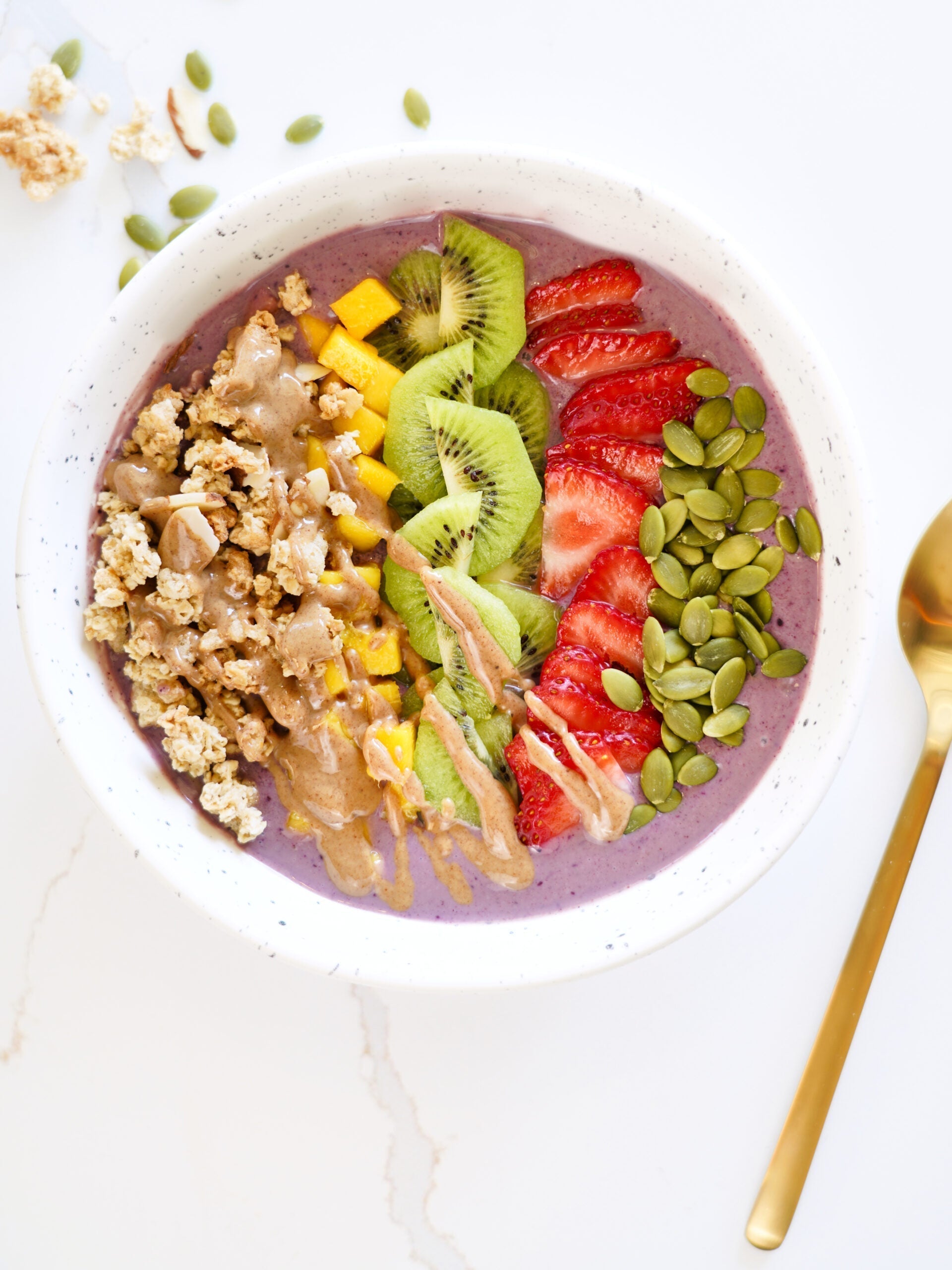 An appetizing açaí bowl with neatly arranged rows of granola, mango, kiwi, strawberries, and pumpkin seeds on a bright, white background.
