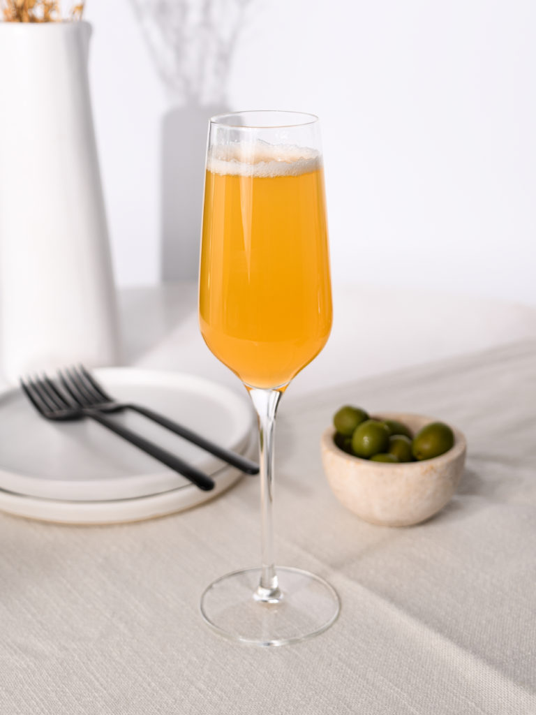 A refreshing mango mimosa in a tall glass, set on a linen tablecloth beside plates and olives for a bright brunch scene. 
