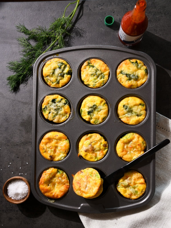 A muffin tin filled with golden egg bites, some with spinach, alongside dill, hot sauce, and salt, creating a savory, appetizing mood.
