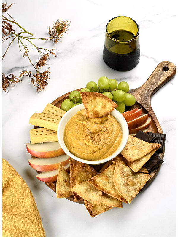 A festive autumnal appetizer: pumpkin pie dip surrounded by cinnamon crisps, grapes, apples, and chocolate on a wooden serving board.
