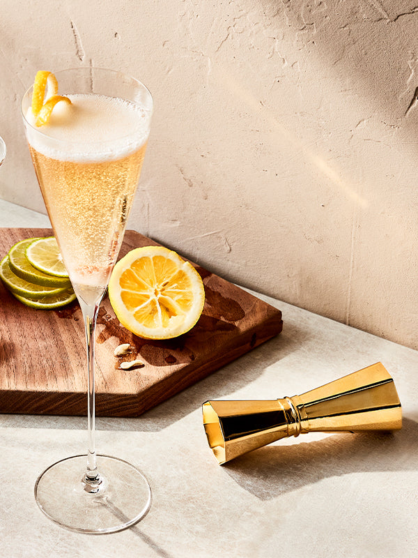 Elegant elderflower champagne cocktail with lemon garnish, golden jigger, and wooden board evoking a celebratory mood.
