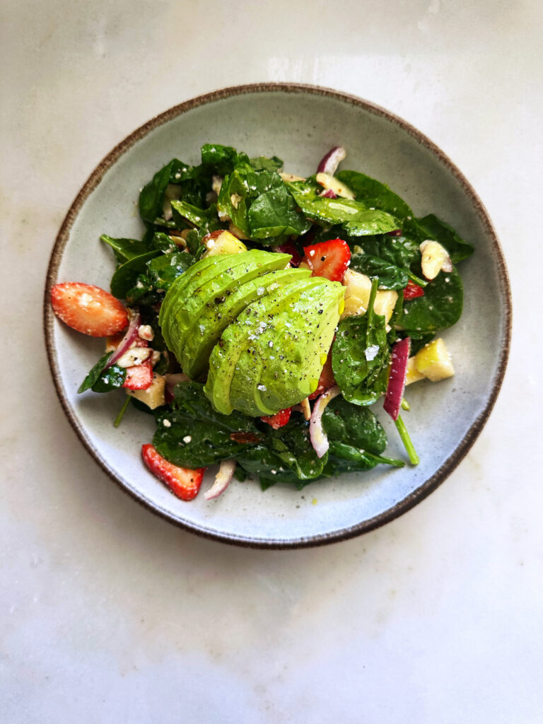An appealing salad with spinach, strawberries, avocado, and red onion in a gray bowl, suggesting a fresh, healthy, and delicious recipe.
