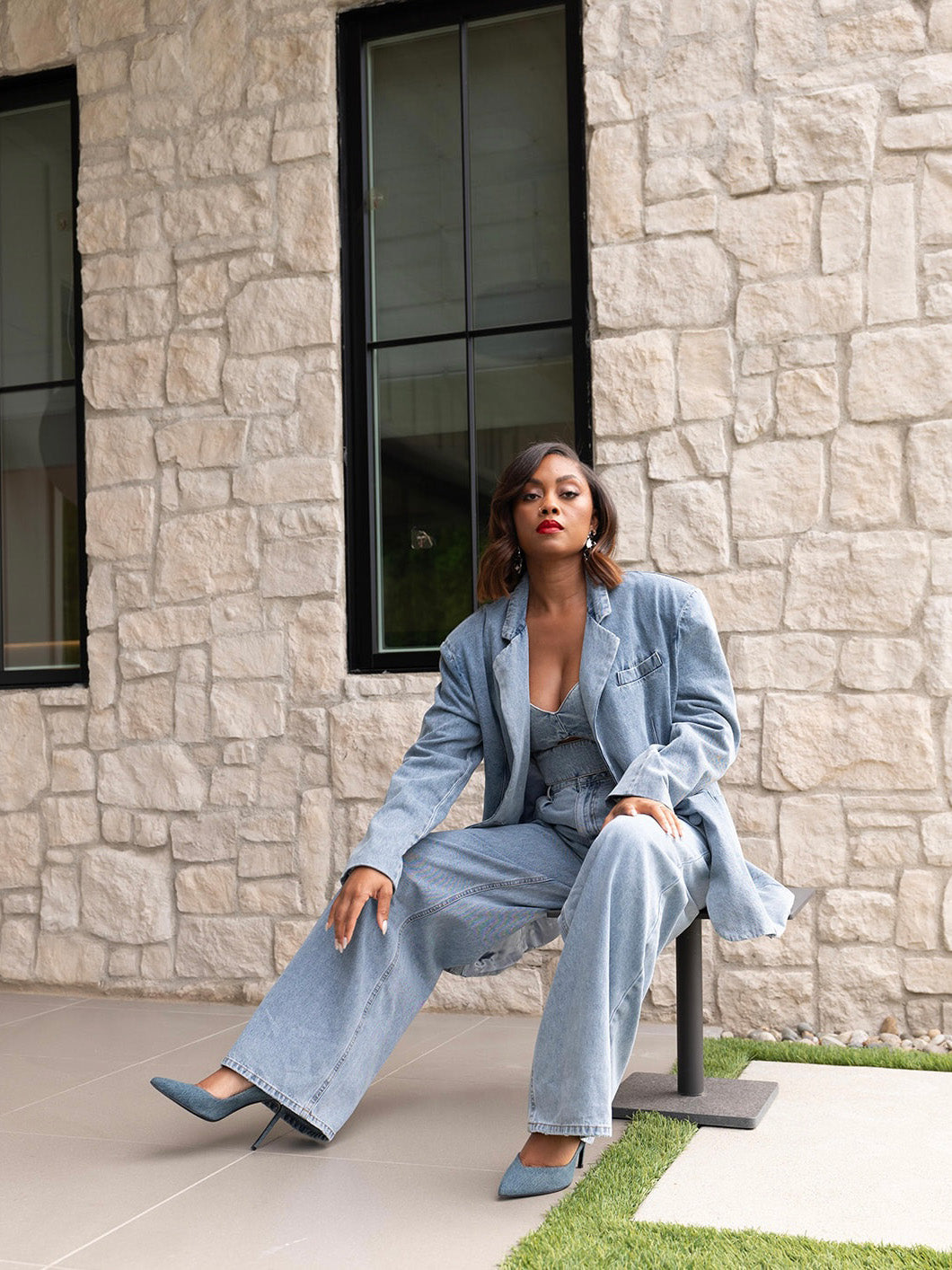 Jada Paul confidently poses in a denim suit, embodying chic sophistication against a textured stone wall and black-framed windows.
