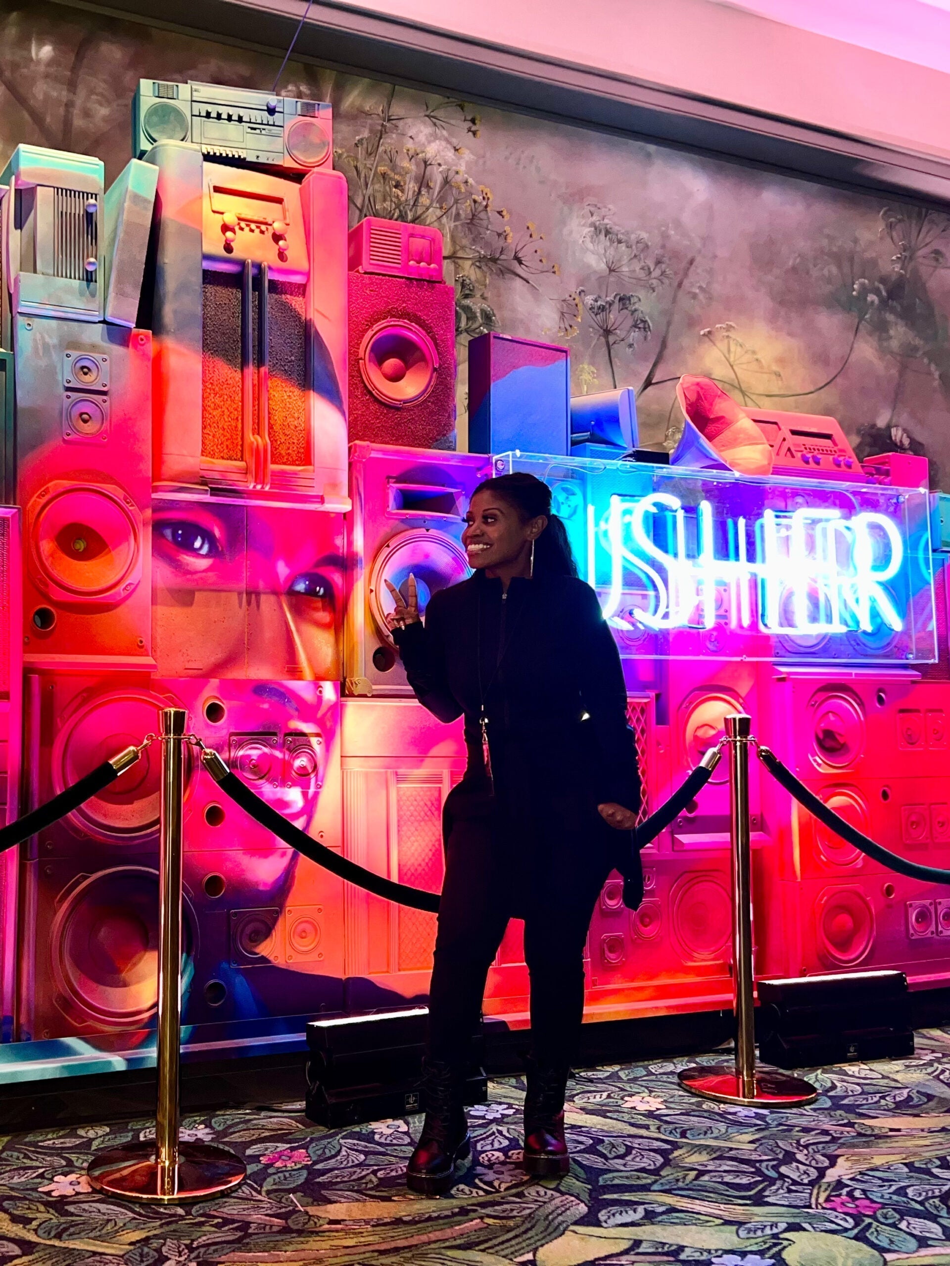 Radiant road manager strikes a pose near neon "Usher" sign, conveying excitement and backstage access to Usher's world.
