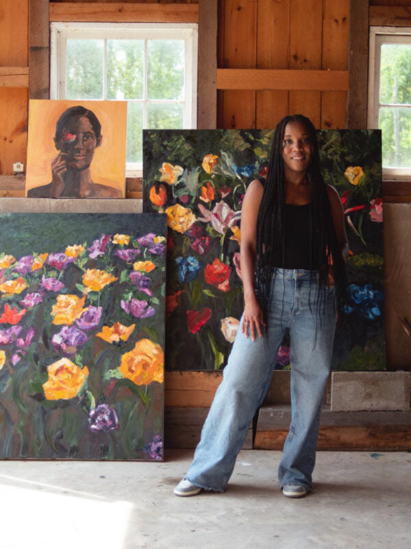A Black female artist smiles confidently amidst her vibrant floral paintings, radiating creativity and empowerment in a rustic studio.
