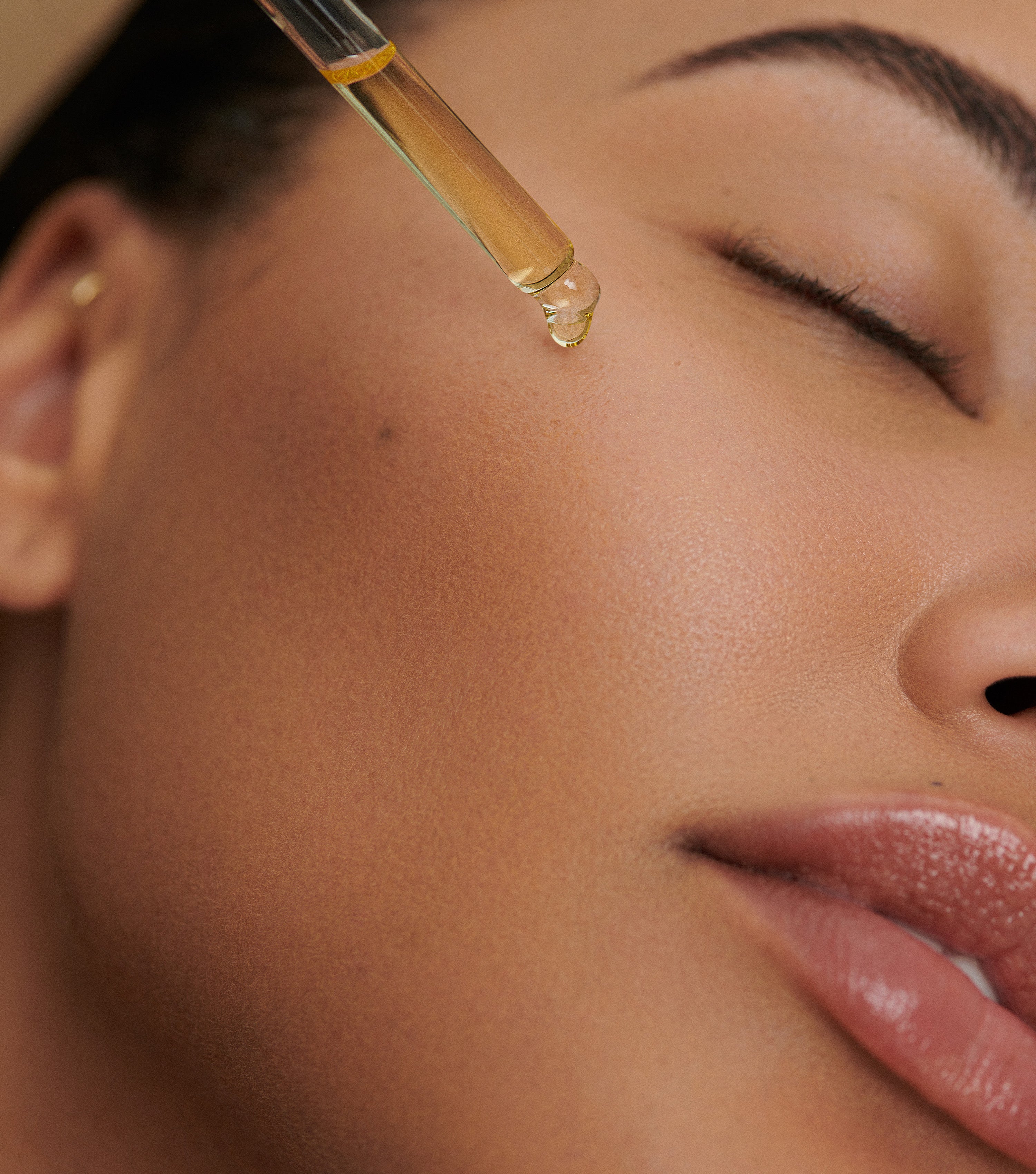 A woman with closed eyes is about to have face oil applied to her cheek, conveying a calming, self-care mood related to skincare.
