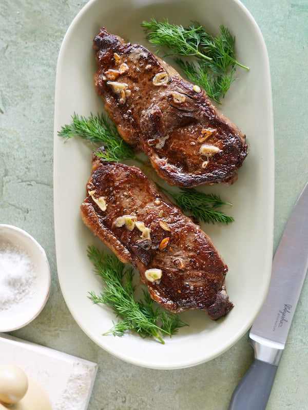 Two pan-seared steaks glistening with garlic butter and fresh dill on a platter, conveying a sense of culinary delight.
