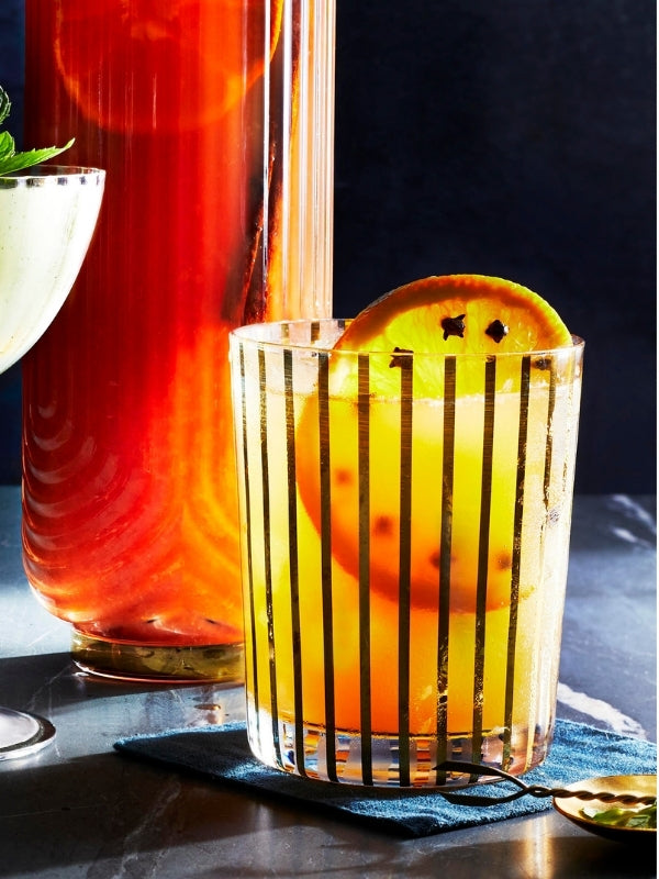 Festive cocktail in a striped glass, garnished with a clove-studded orange slice, next to a tall glass pitcher.
