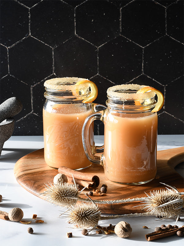 Two mason jars of smoky citrus cider, garnished with orange peels, evoke a cozy, autumnal mood. Spices and thistles add rustic charm.
