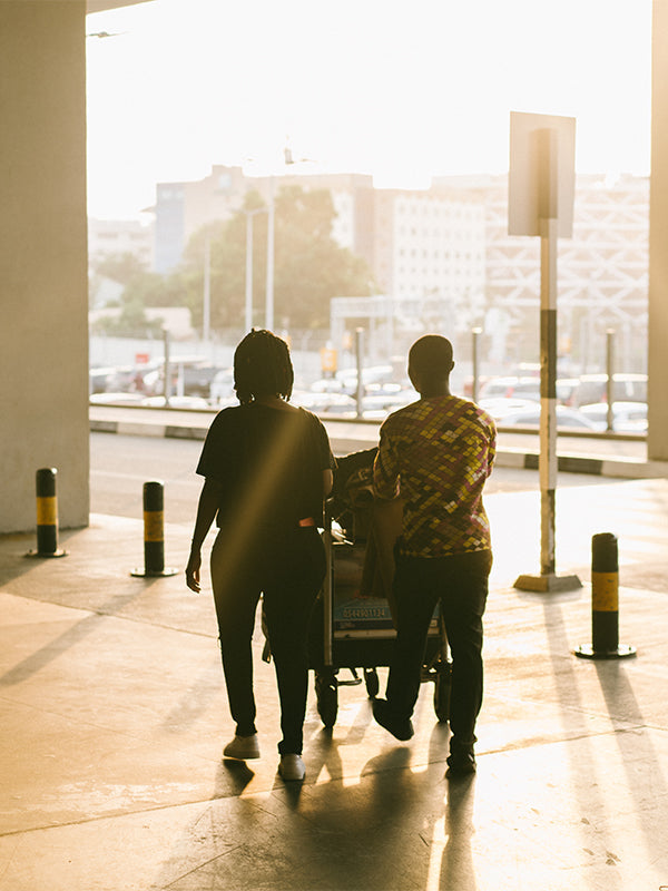 A couple wheels a luggage cart toward the airport exit, silhouetted against the bright sunlight, hinting at a hopeful journey together.
