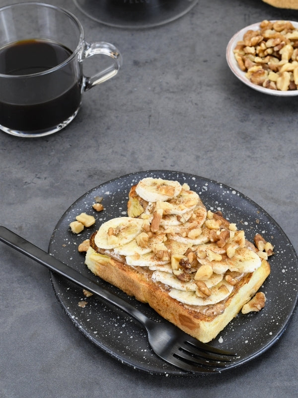 Close-up of banana bread toast with sliced bananas and walnuts on a speckled plate, conveying a cozy breakfast vibe. 
