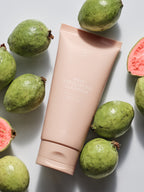 Peach-colored exfoliating cleanser by Sweet July Skin, surrounded by guavas on a white background.
