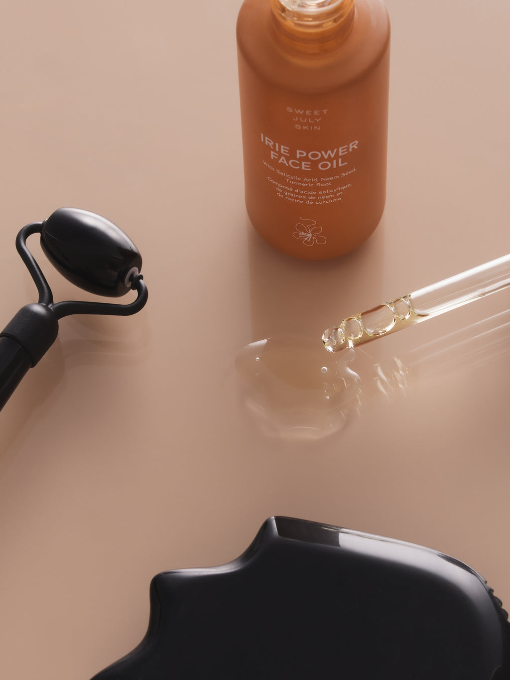 Bottle of face oil by Sweet July Skin, alongside a black facial roller, serum, and gua sha tool.