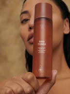 Brown glass bottle of Pava Toner by Sweet July Skin, held in hand against a blurred face.
