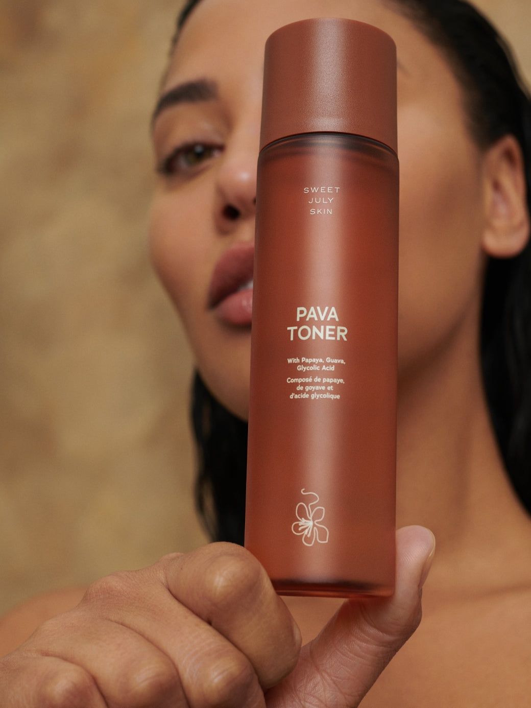 Brown glass bottle of Pava Toner by Sweet July Skin, held in hand against a blurred face.
