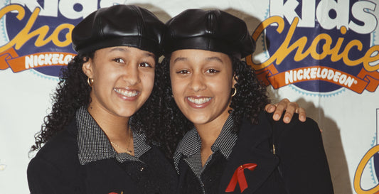 The image features two young girls hugging and smiling are indoors, posing against a wall, and wearing fashionable clothing and accessories.