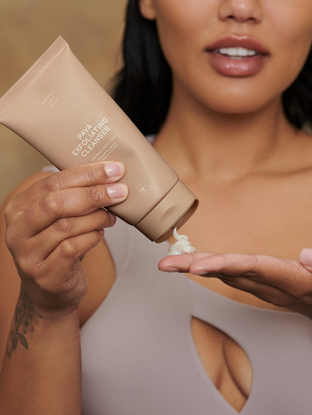 Tan exfoliating cleanser by Sweet July Skin being squeezed onto a hand, with a woman's upper torso in the background.
