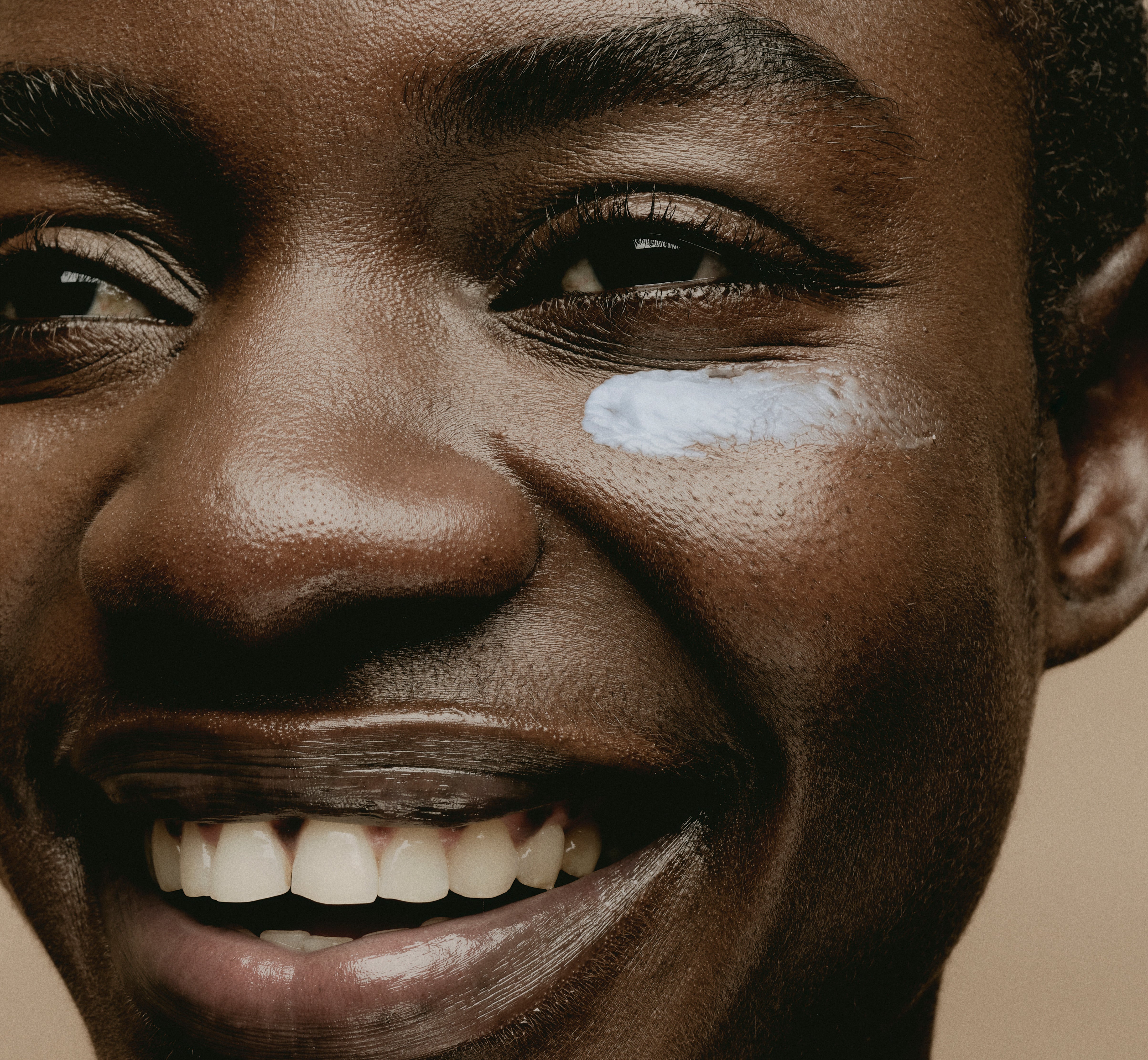 Close up of model smiling with Sweet July Skin Coffee Fix Peptide Eye Cream swatch under left eye displaying the light blue hue.  
