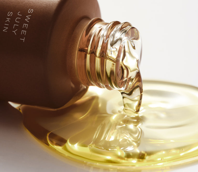Close-up of a tilted brown bottle with "SWEET JULY SKIN" text, pouring golden oil onto a white surface. Fluid dynamics and reflections create a glistening, glossy effect.