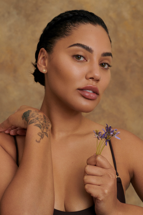 Close-up of a woman with braided hair, holding purple flowers, showcasing a tattoo on her arm, against a tan background.