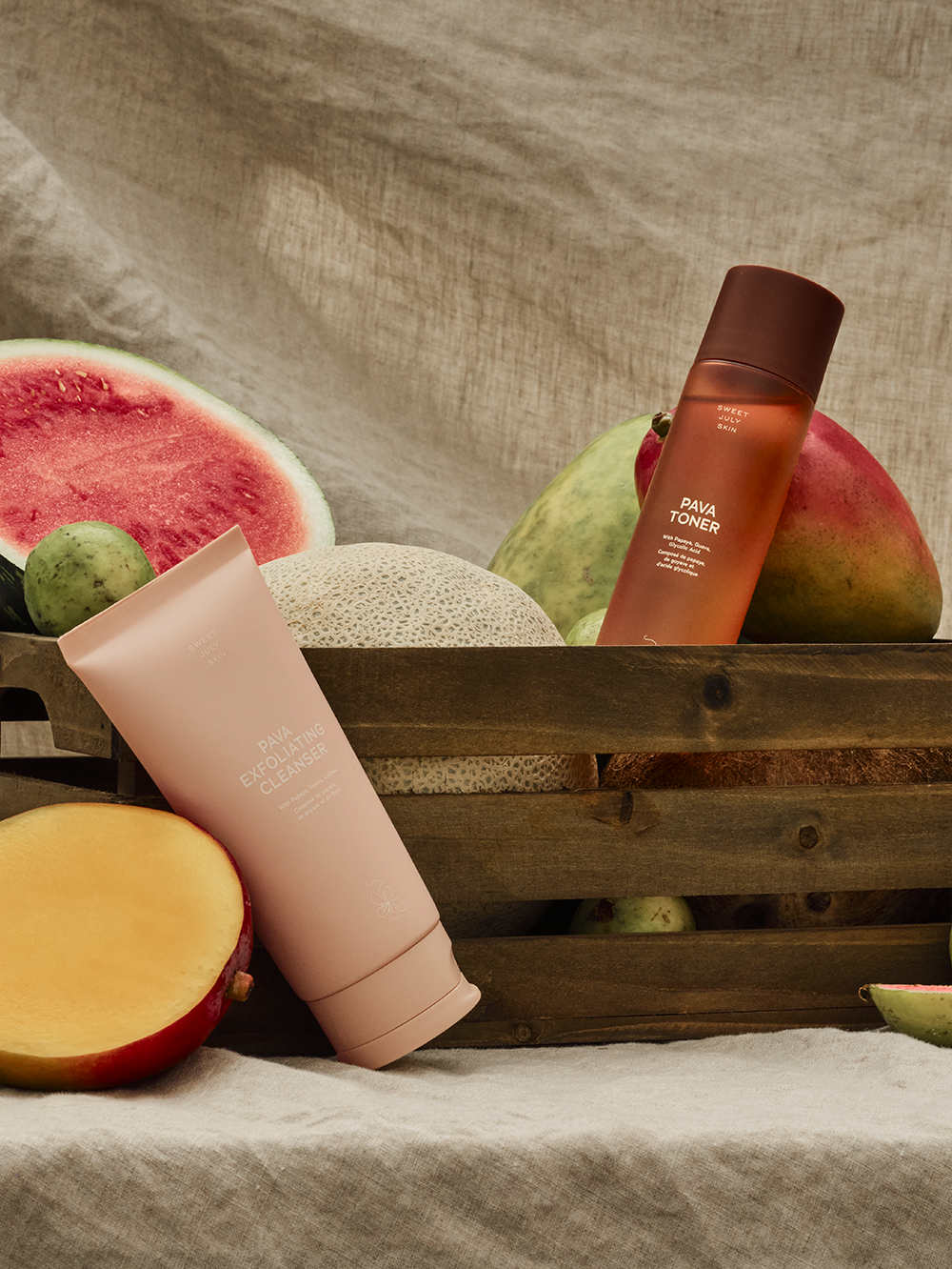 Skincare duo by Sweet July Skin featuring a toner and exfoliating cleanser surrounded by fruit in a wooden crate.
