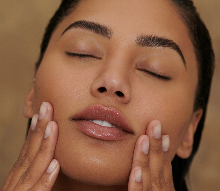 A woman with tan skin and wet, dark hair touches her face with her fingers, eyes closed in a relaxed expression. Her lips are glossy and slightly parted.