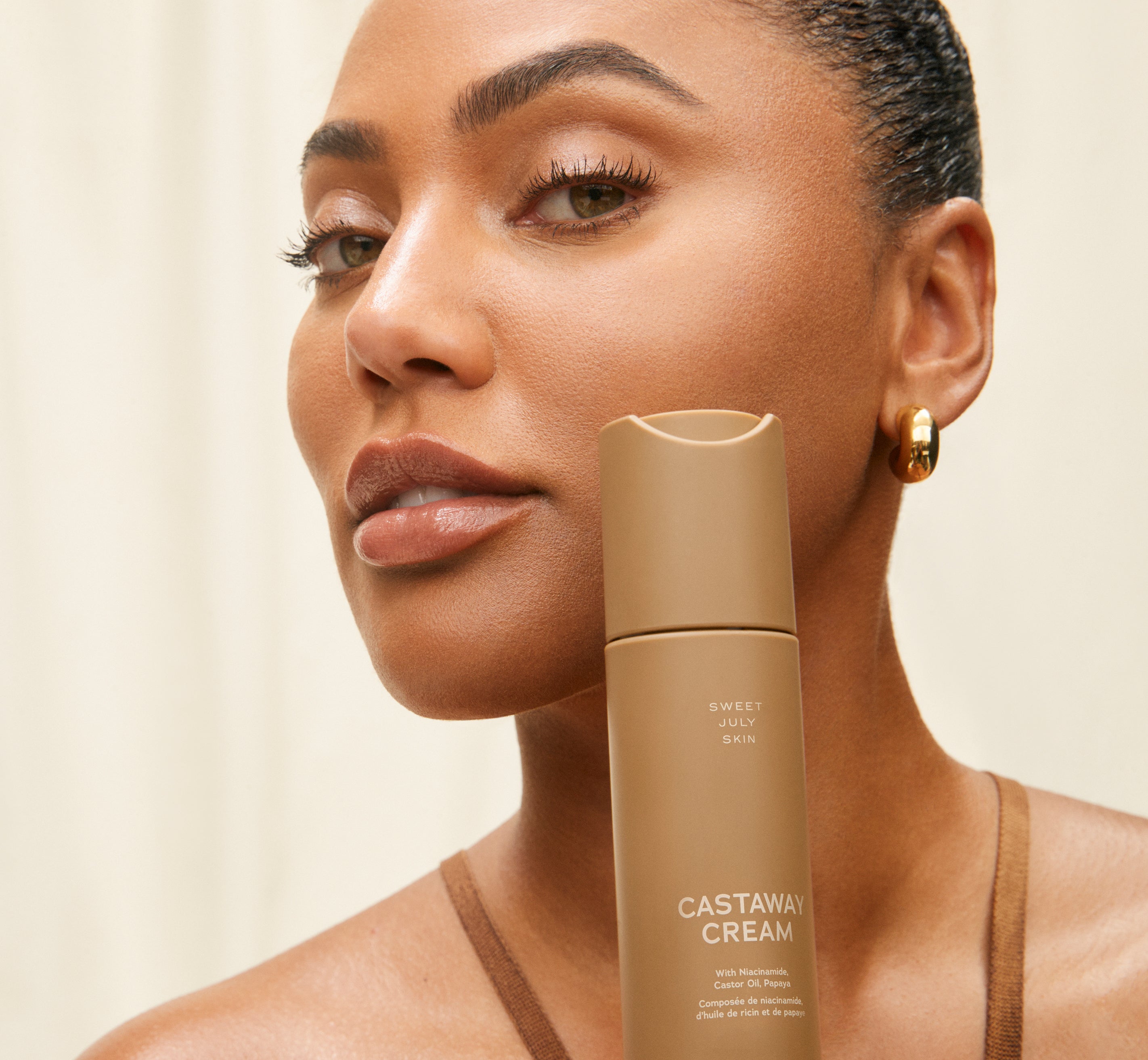 Sweet July founder Ayesha Curry holding a bottle of Castaway Cream against her cheek, before a cream curtain.