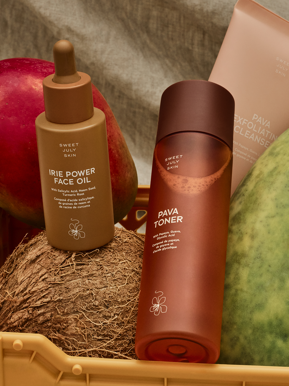 Skincare set by Sweet July Skin, featuring a face oil, a toner, and an exfoliating cleanser alongside tropical fruits and a coconut.
