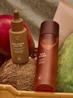 Skincare set by Sweet July Skin, featuring a face oil, a toner, and an exfoliating cleanser alongside tropical fruits and a coconut.
