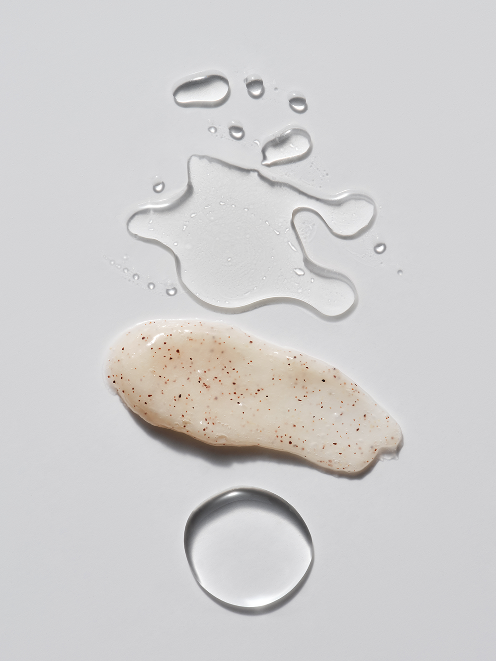 Facial scrub swatch by The Essentials Set with exfoliating particles, alongside clear liquid droplets on a white surface.
