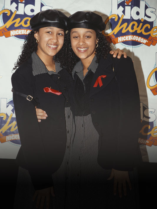 The image features two young girls hugging and smiling are indoors, posing against a wall, and wearing fashionable clothing and accessories.